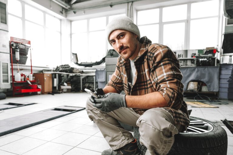 Car service worker sitting on a wheel and resting, using mobile phone